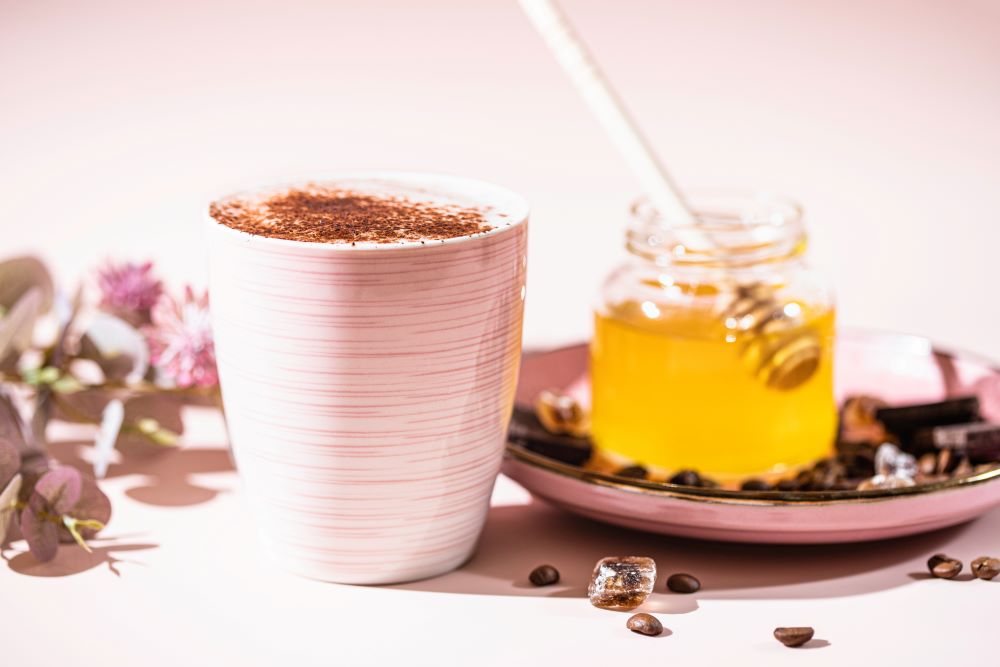 A hot latte and iced latte with whipped cream beside a bag of Nummy Creations Vanilla herbal coffee alternative.